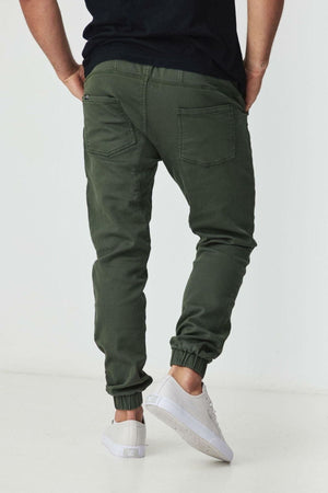 A person wearing BLKWD James Jogger Pants in dark green cotton stretch, a black t-shirt, and white sneakers stands with their back to the camera against a plain white background.