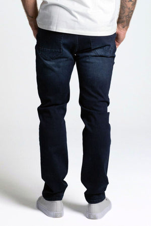 Someone wearing a white T-shirt, BLKWD West Denim Jean Slim in a 5-pocket style, and gray sneakers stands with their back to the camera, hands in pockets, against a plain light background.