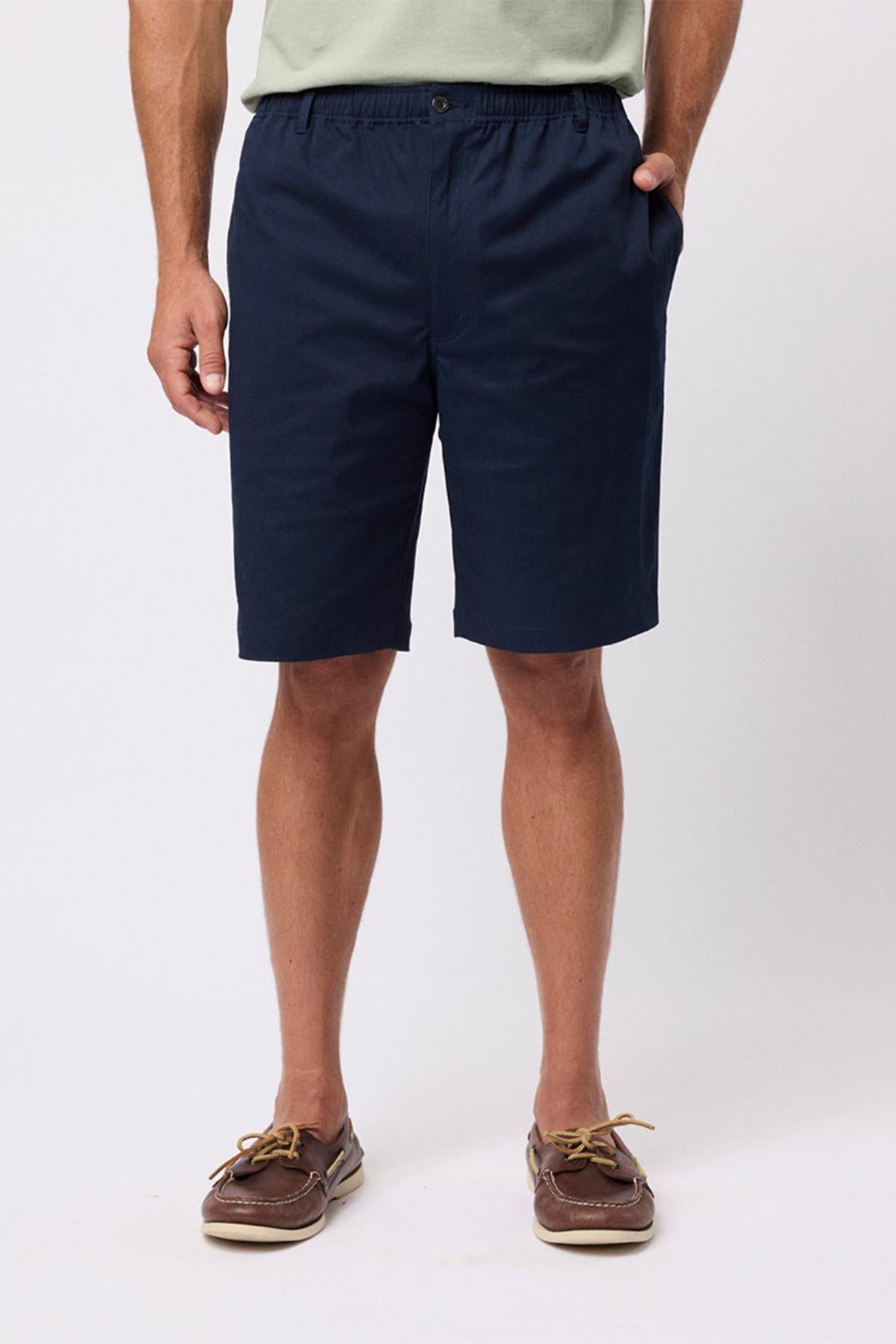 A person wearing navy blue Breakaway Cody Shorts with an elastic waistband, a light green shirt, and brown boat shoes stands hands in pockets against a plain white backgroundβperfect for everyday style.