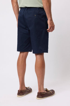 A man is shown from behind wearing a light green shirt, brown boat shoes, and navy blue Breakaway Cody Shorts with an elastic waistband. The focus on his lower body highlights the everyday comfort of these shorts against a plain background.