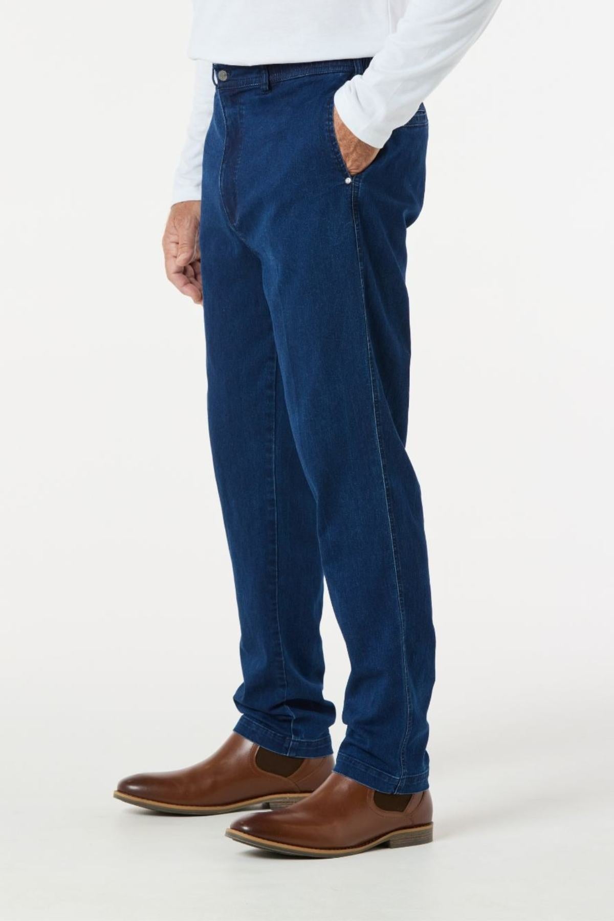 A person is showcasing the Breakaway Intense Denim Jean in dark blue, paired with a white long-sleeve shirt and brown leather boots. They are posed against a plain white background with one hand casually resting in their pocket, drawing attention to the clothing ensemble.