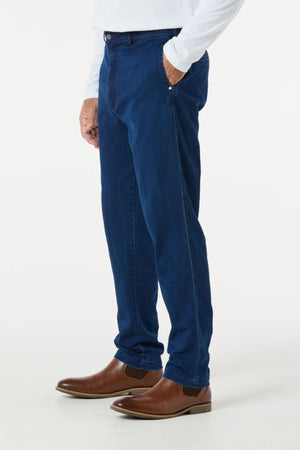 A person is showcasing the Breakaway Intense Denim Jean in dark blue, paired with a white long-sleeve shirt and brown leather boots. They are posed against a plain white background with one hand casually resting in their pocket, drawing attention to the clothing ensemble.