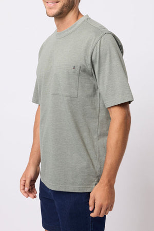 A man in a relaxed fit, light gray-green Breakaway Kinnersly Crew T-shirt with a chest pocket and navy blue shorts stands against a plain white background, smiling slightly.