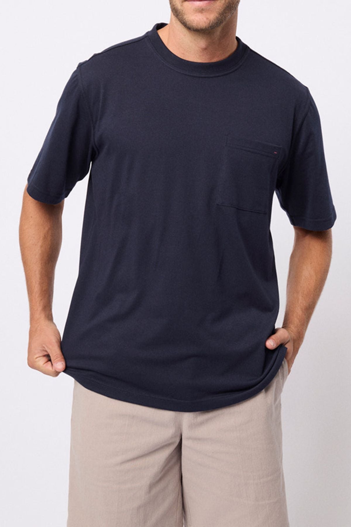 A man wears the Breakaway Kinnersly Crew in dark navy with a chest pocket and light beige shorts, standing against a plain white background and holding the shirt’s hem. Its relaxed fit and lightweight fabric offer all-day comfort.