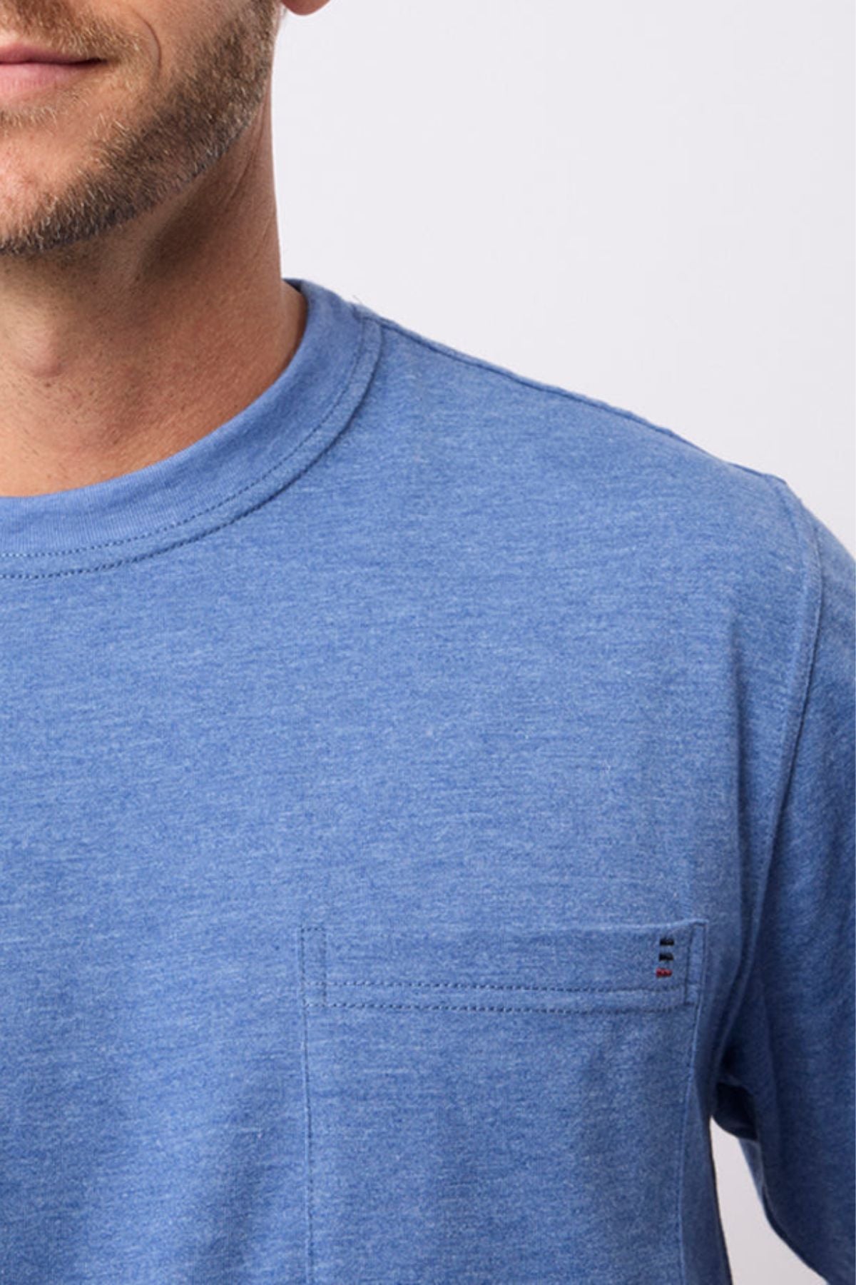 A close-up shows a man in the Breakaway Kinnersly Crew, a relaxed-fit blue tee with a chest pocket. The image features only his lower face, neck, and upper torso set against a plain light background.