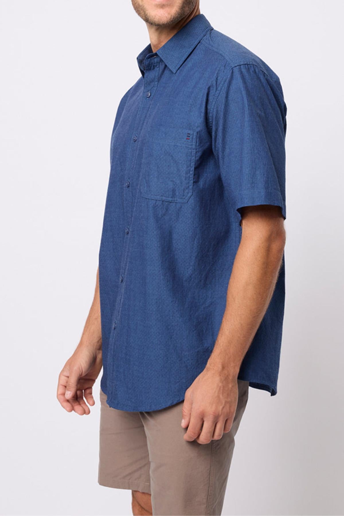 A man in the Breakaway Olivero Shirt—a short-sleeve, blue 100% cotton men’s shirt with a chest pocket—paired with khaki shorts, stands against a plain light background.
