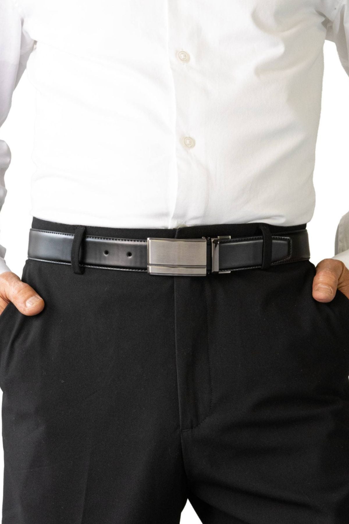 A person wears a white dress shirt tucked into black dress pants with the Buckle 5519 Heritage 35mm Reversible Leather Belt, featuring a silver rectangular buckle, and stands with hands in their pockets.
