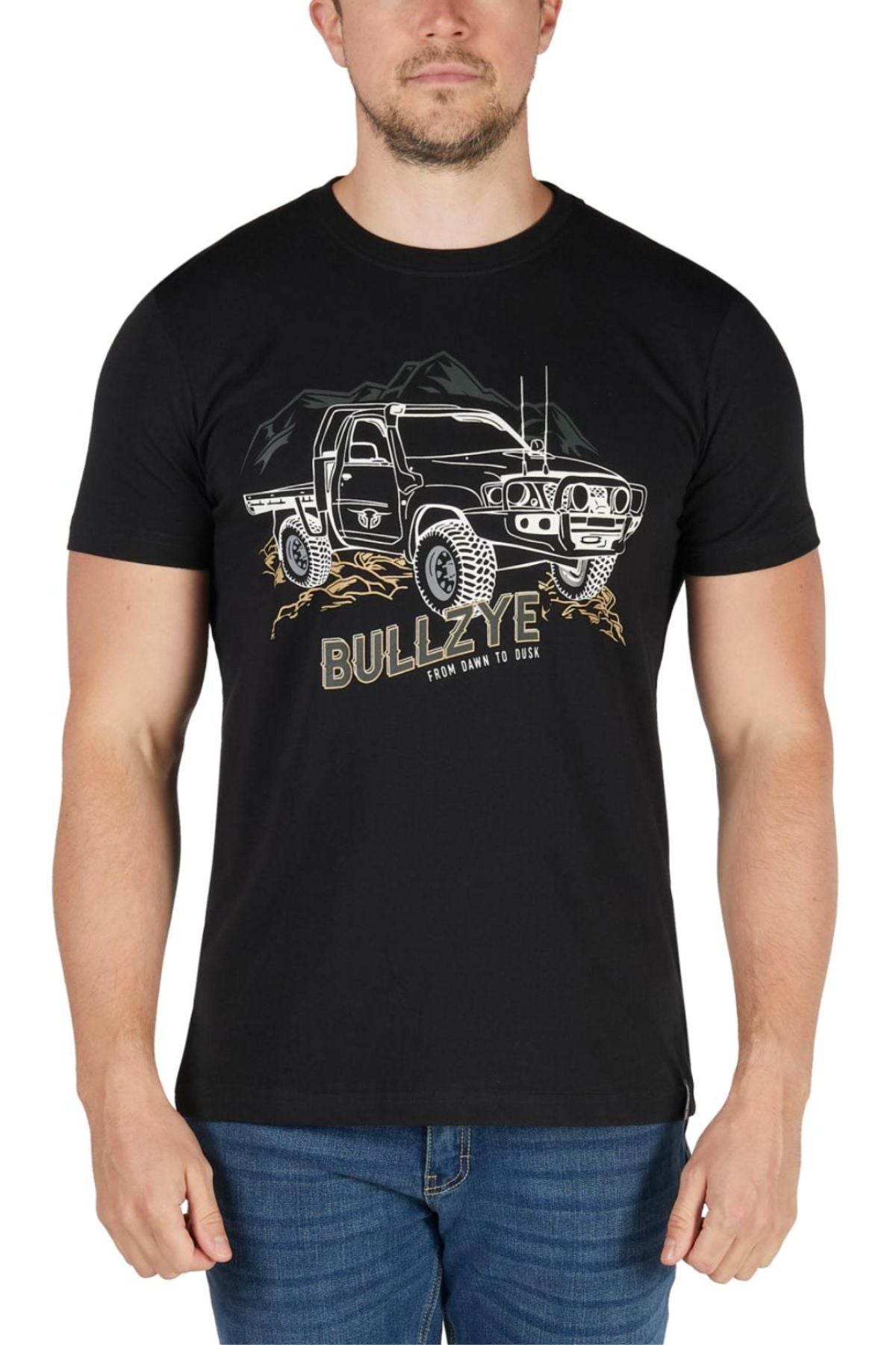 A man wears the Bullzye Dusk Tee, featuring an illustrated off-road truck, mountains, and "FROM DAWN TO DUSK" on the front. Made from soft cotton jersey, this classic tee pairs perfectly with his blue jeans.