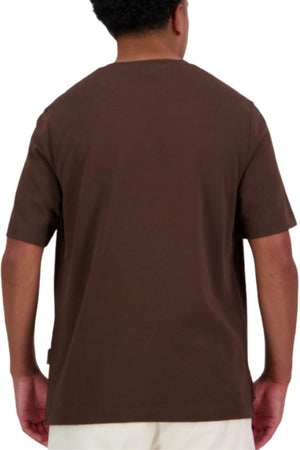 A person with short curly hair is seen from behind wearing an oversized brown Canterbury CCC Collection T-Shirt and light-colored pants against a white background.