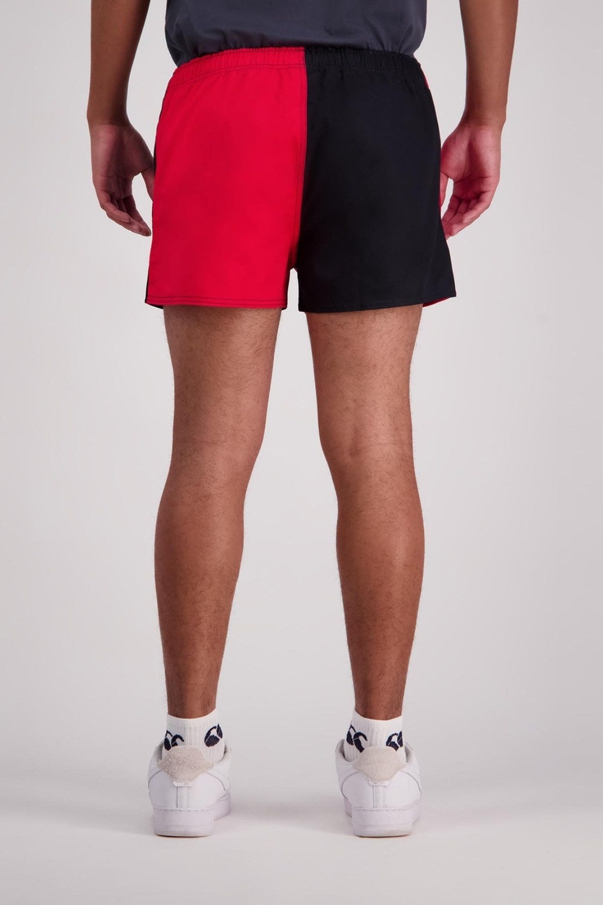 A person dressed in Canterbury Cotton Twill Harlequin Shorts, featuring a half red and half black design, paired with white socks and sneakers. The image captures them from behind, highlighting the elasticated waist of the shorts against a plain background.
