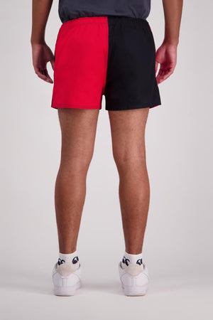 A person dressed in Canterbury Cotton Twill Harlequin Shorts, featuring a half red and half black design, paired with white socks and sneakers. The image captures them from behind, highlighting the elasticated waist of the shorts against a plain background.