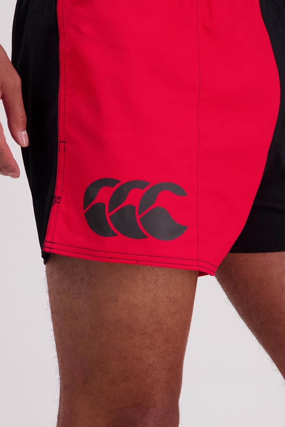 A close-up view of a person wearing the Canterbury Cotton Twill Harlequin Short, featuring red and black elasticated waist shorts with a distinctive triple "C" logo on the left leg. These breathable cotton shorts showcase a middle seam in the red section, set against a simple light gray background.