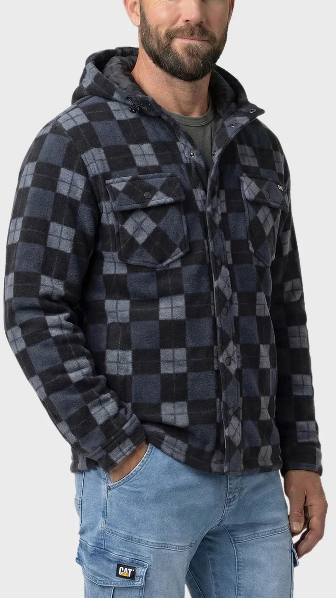A man stands against a light gray background wearing a black and gray plaid CAT Active Work Jacket with an insulated hood over a dark shirt and light blue CAT work pants, with one hand in his pocket.