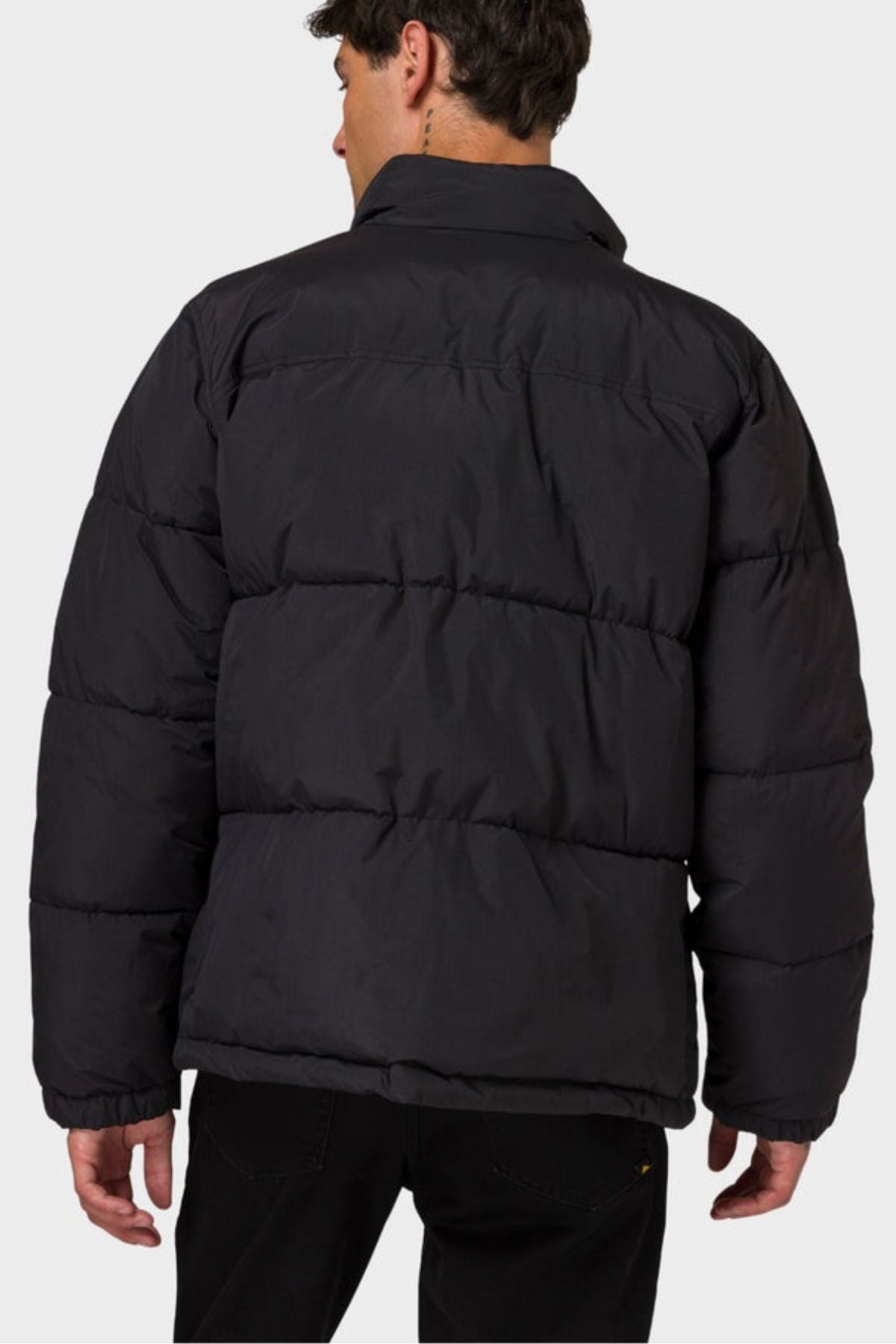 A person is shown from behind, looking slightly to the side against a plain light gray background, wearing the CAT Heavy Weight Insulated Puffer Jacket and black pants.