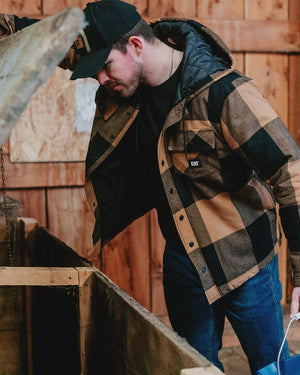 A man in a CAT Hooded Flannel Shirt Jacket with recycled insulation and a black cap lifts the lid of a wooden container inside a rustic hut.