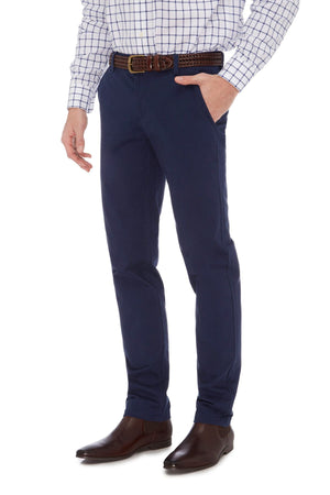 A person is standing with one hand in their pocket, wearing a white checkered shirt, dark blue City Club Hammond Place pants made of stretch cotton, a brown woven leather belt, and brown dress shoes. The slim-fit design of the pants elegantly highlights their silhouette. The person is visible from the neck down.