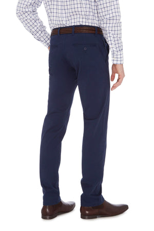 A person wearing a checkered long-sleeve shirt, City Club Hammond Place Pant, a brown braided belt, and brown dress shoes is standing with their back facing the camera. The person's left arm is bent, resting on their hip.