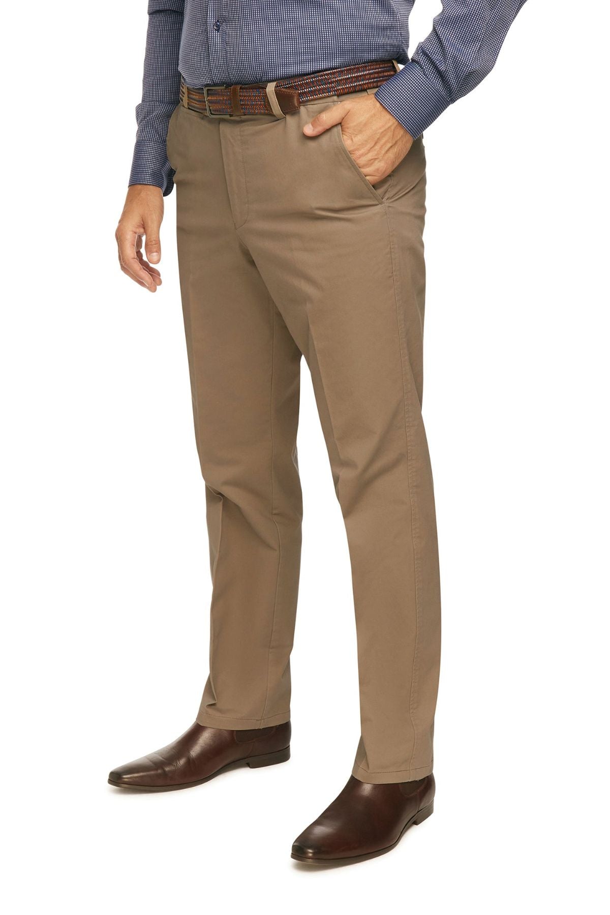 A person is standing with one hand in their pocket, wearing a blue long-sleeve shirt, brown belt, City Club Mariner Harbour Pants in beige featuring elasticated sides for comfort, and brown dress shoes. The image focuses on their lower body, from mid-chest downward.
