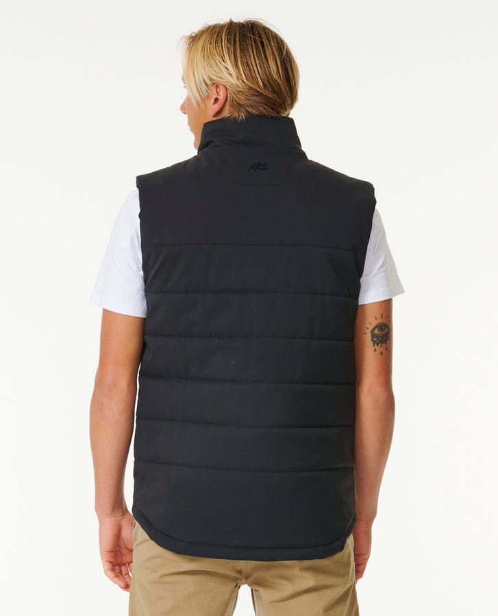A person with blonde hair, viewed from the back, is donned in a black Rip Curl Ridge Anti Series Vest layered over a white T-shirt and beige pants. A tattoo is visible on their left arm. The backdrop is plain white. This winter apparel item provides durable water-resistant protection against cold weather.