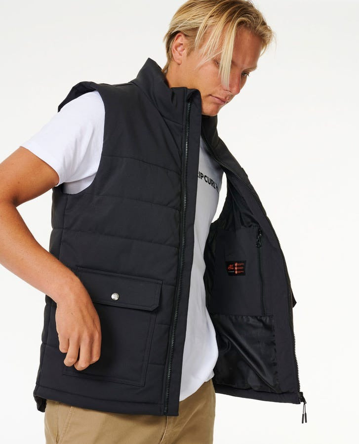 A person with casually styled blonde hair, sporting a white t-shirt and khaki pants, is showcasing the inner lining of the black Rip Curl Ridge Anti Series Vest. This durable water-resistant vest includes a zippered closure and a buttoned side pocket. The individual’s gaze is directed downward, making it an ideal choice for winter attire.