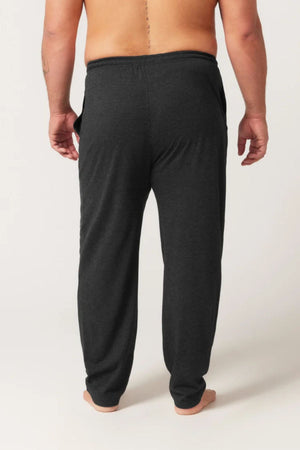 A person stands barefoot with their back to the camera, wearing dark gray Contare Country Bamboo Cotton Lounge Pants. The background is plain and light-colored.