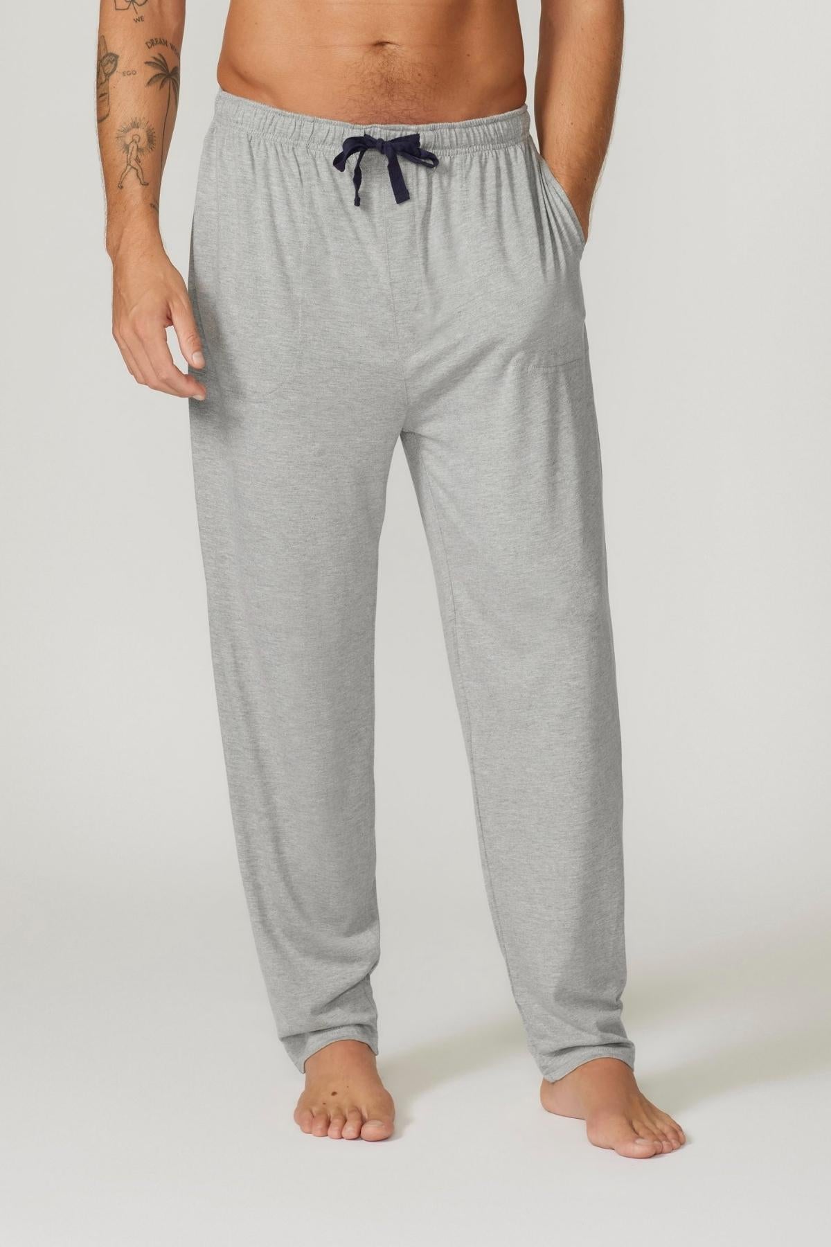 A person stands barefoot wearing Contare Country Bamboo Cotton Lounge Pants in light gray, featuring a relaxed fit and an adjustable drawstring waistband. Their right arm, adorned with tattoos, is partially visible. The lounge pants include side pockets. The background is plain and light-colored.