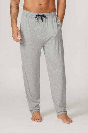 A person stands barefoot wearing Contare Country Bamboo Cotton Lounge Pants in light gray, featuring a relaxed fit and an adjustable drawstring waistband. Their right arm, adorned with tattoos, is partially visible. The lounge pants include side pockets. The background is plain and light-colored.