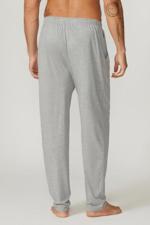 A person standing barefoot, facing away from the camera, wearing Contare Country Bamboo Cotton Lounge Pants in light gray. Their left arm, adorned with tattoos on the arm and shoulder, hangs by their side. The lounge pants are loose-fitting and appear to have pockets on both sides.