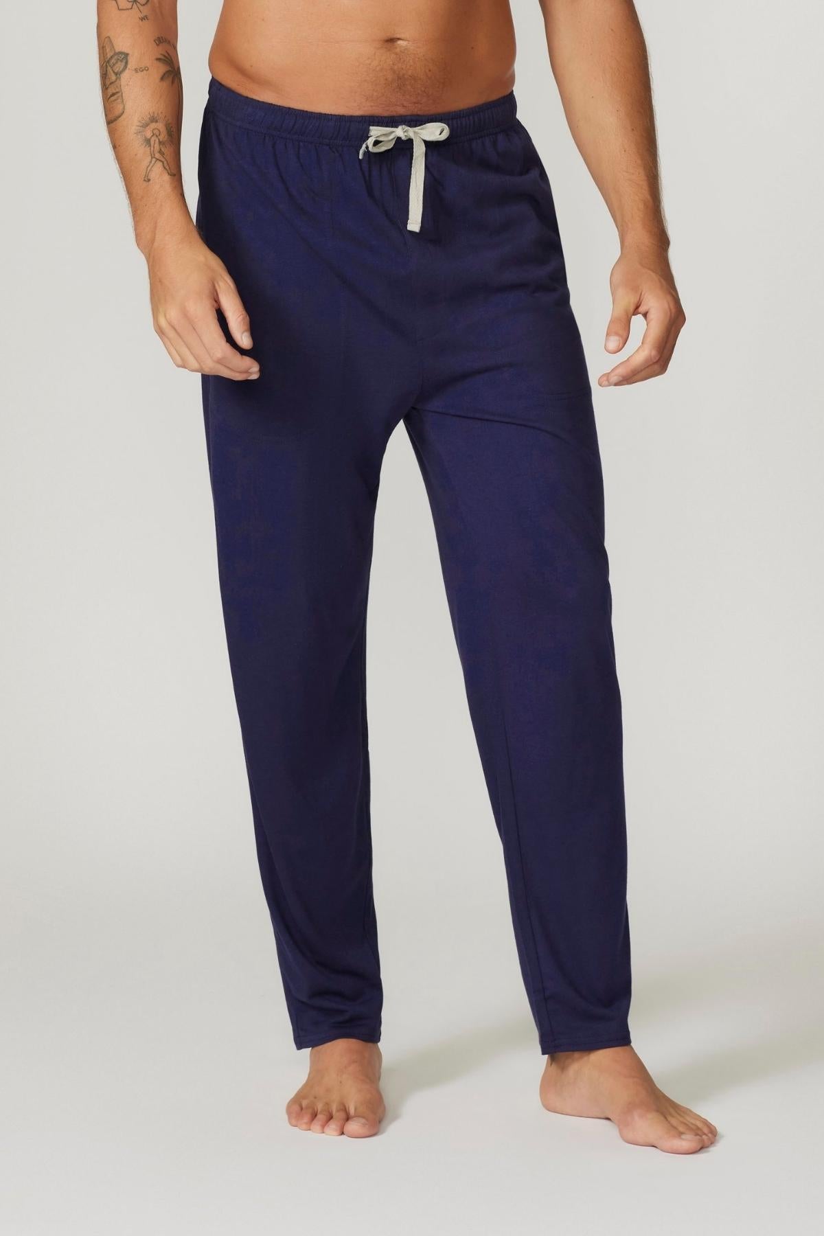 A man stands barefoot in Contare Country Bamboo Cotton Lounge Pant, navy blue with a light gray drawstring. The breathable fabric enhances his casual style. His tattooed left arm is partly visible against a plain light gray background.