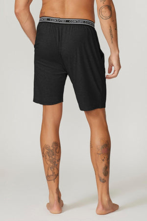 A person with visible tattoos on their arms and legs is standing barefoot, facing away. They are wearing Contare Country Bamboo Cotton Sleep Shorts in black, which feature an elastic waistband with a repeating "CONTRARY" text. Their hands are resting casually at their sides.