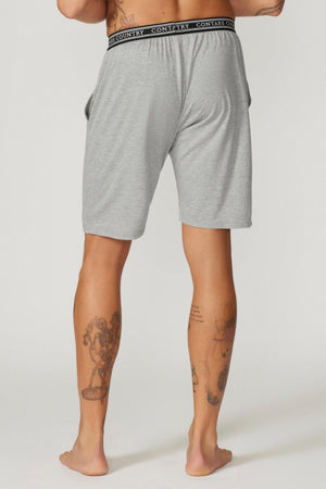 A person stands facing away from the camera, barefoot and revealing tattoos on their left calf and right thigh. They are wearing light gray Contare Country Bamboo Cotton Sleep Shorts, which feature a black waistband repeatedly printed with the text "Contrary.