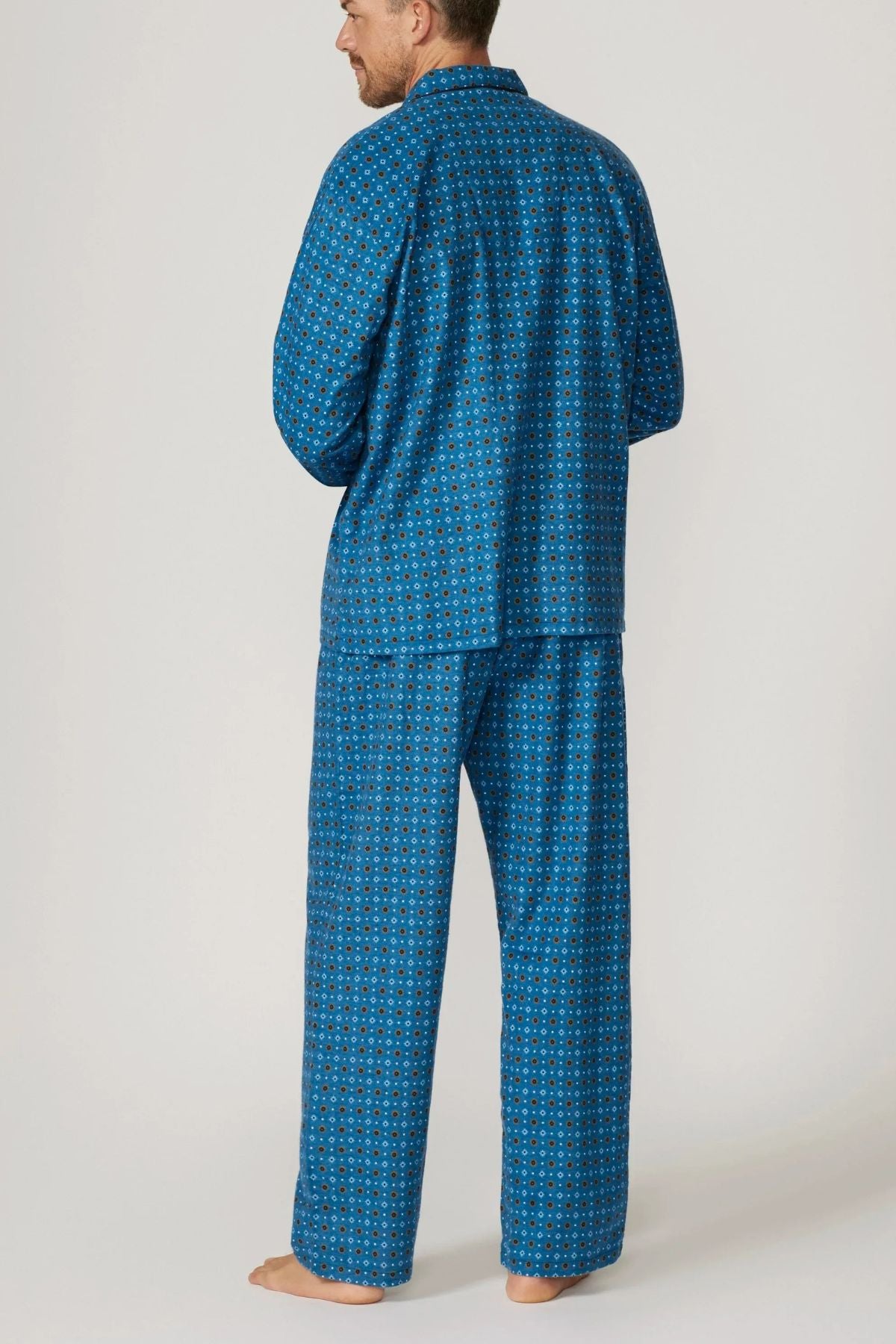 A man stands barefoot, facing away, wearing the Contare Country Winter Flannelette PJ Set—a blue brushed cotton pyjama with a small geometric print. The long-sleeve top and loose pants offer comfort against a light plain background.