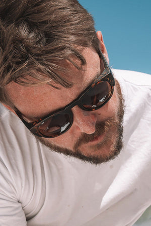 A man with brown hair and a beard, wearing SafeStyle Cruisers Matte Brown Tort sunglasses with brown polarised lenses and a white shirt, looks downward under bright sunlight against a clear blue sky.