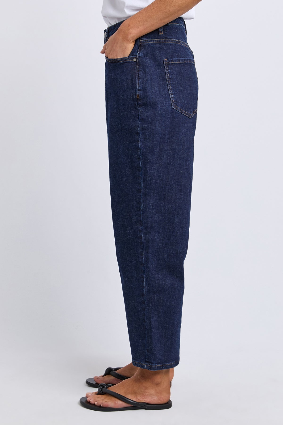 A person stands in profile against a plain light background wearing Foxwood Ambrose Barrel Jean, a dark blue high-waisted stretch denim, with one hand in their pocket and black sandals.