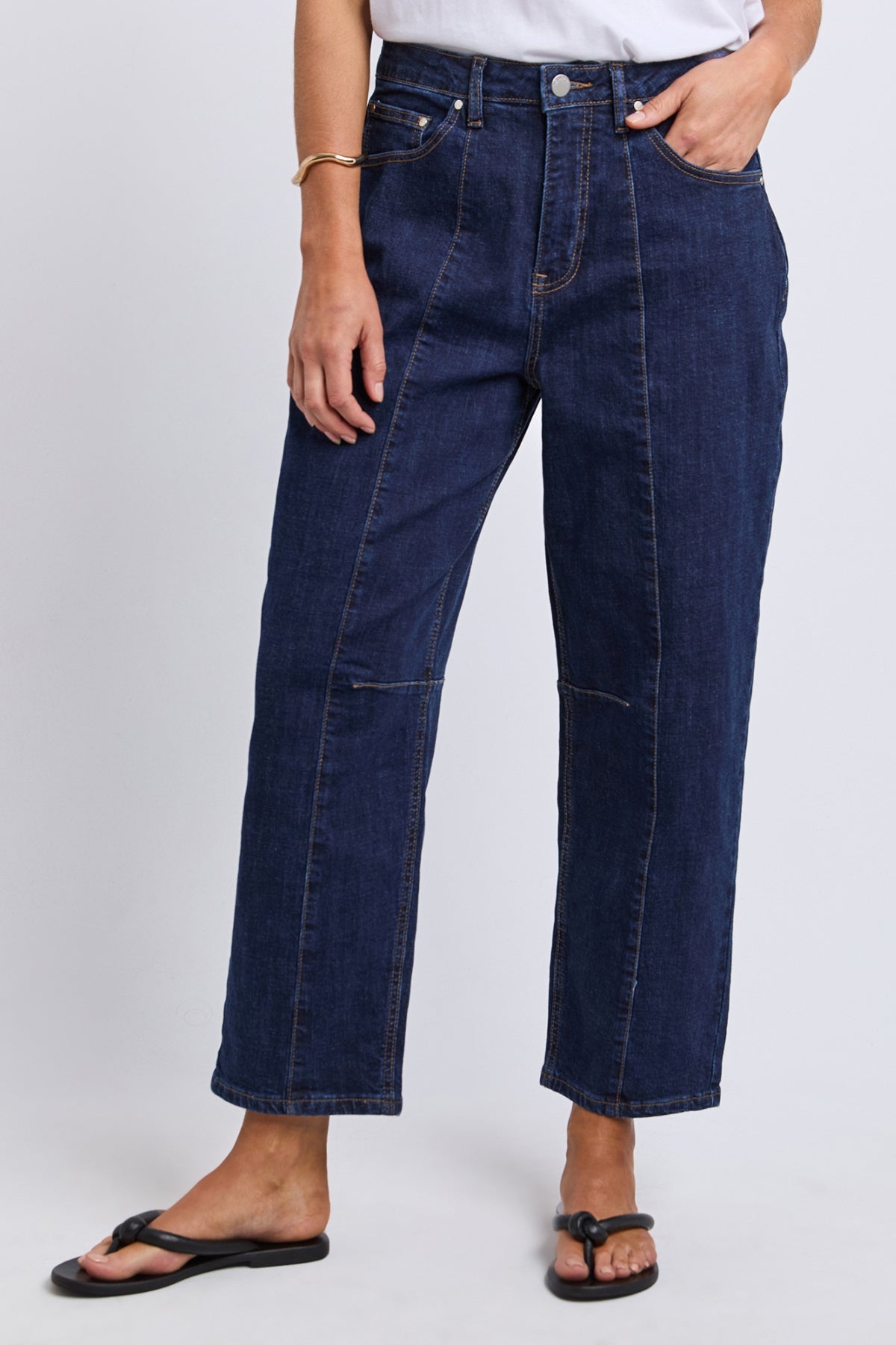 A person wearing Foxwood Ambrose Barrel Jean in dark blue, styled with a white shirt, bracelet, and black sandals, stands against a plain light background.