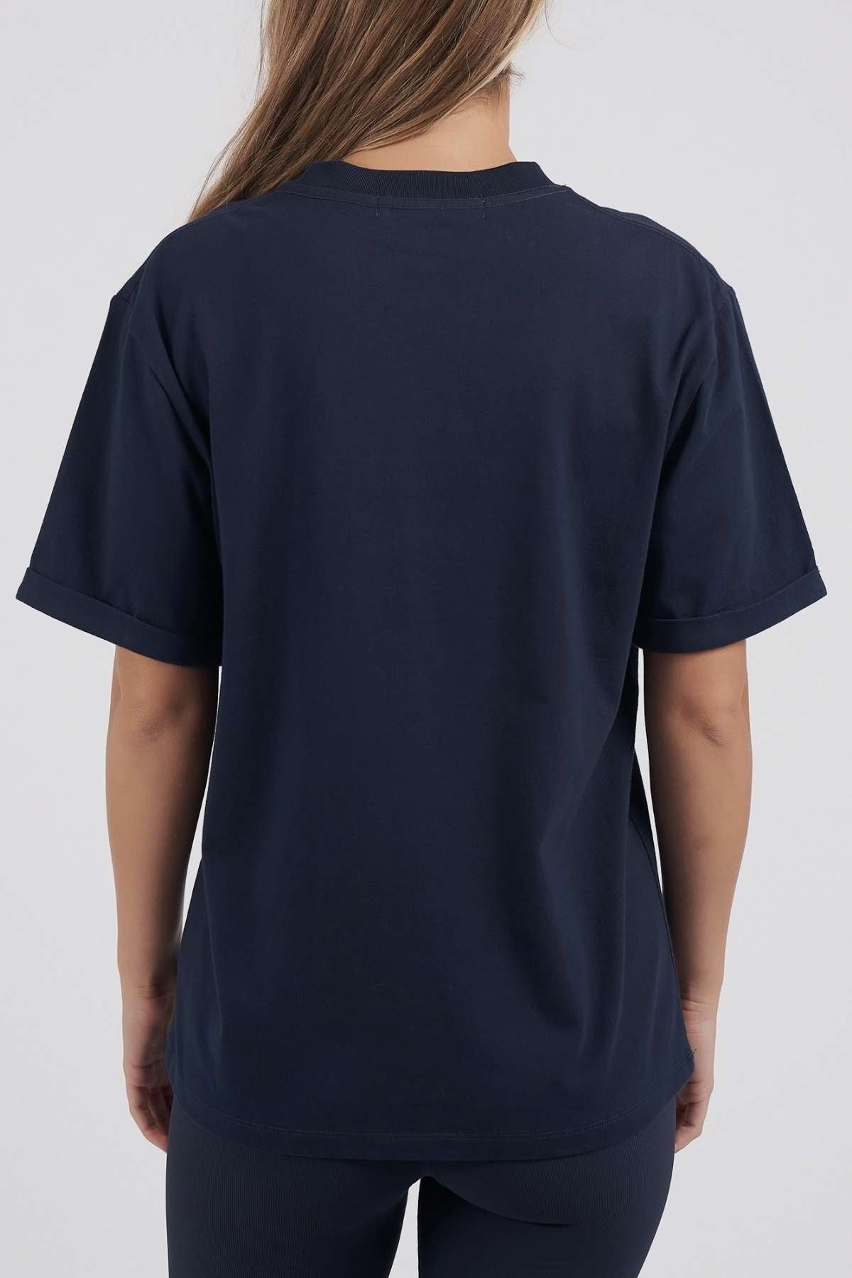 A person with long hair wears a Foxwood Athletics Tee in plain dark blue, facing away from the camera against a neutral backdrop, highlighting the leisurewear's subtle embossed chest artwork.
