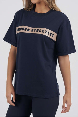 A woman wears a Foxwood Athletics Tee in navy with embossed chest artwork in a beige band. She pairs it with dark pants, making this leisurewear pop against the plain white background.