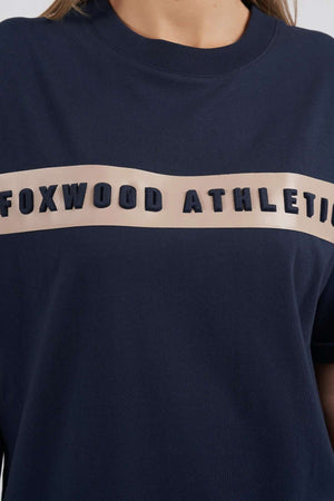 A close-up of the Foxwood Athletics Tee shows its embossed chest artwork on a beige band. Modeled by a person with long, light brown hair, this stylish leisurewear highlights both comfort and design.