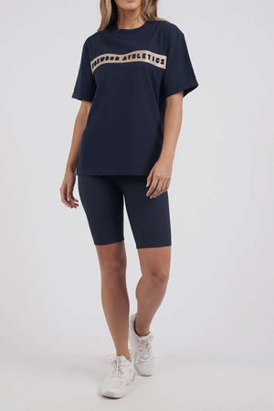 A person sports the Foxwood Athletics Tee, a navy leisurewear piece featuring embossed chest artwork, paired with matching bike shorts and crisp white sneakers, against a plain white backdrop.