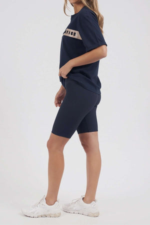 Sporting a navy blue Foxwood Athletics Tee with embossed chest artwork, matched with navy cycling shorts and white sneakers, a person stands sideways facing right against a plain white backdrop—ideal for stylish leisurewear enthusiasts.