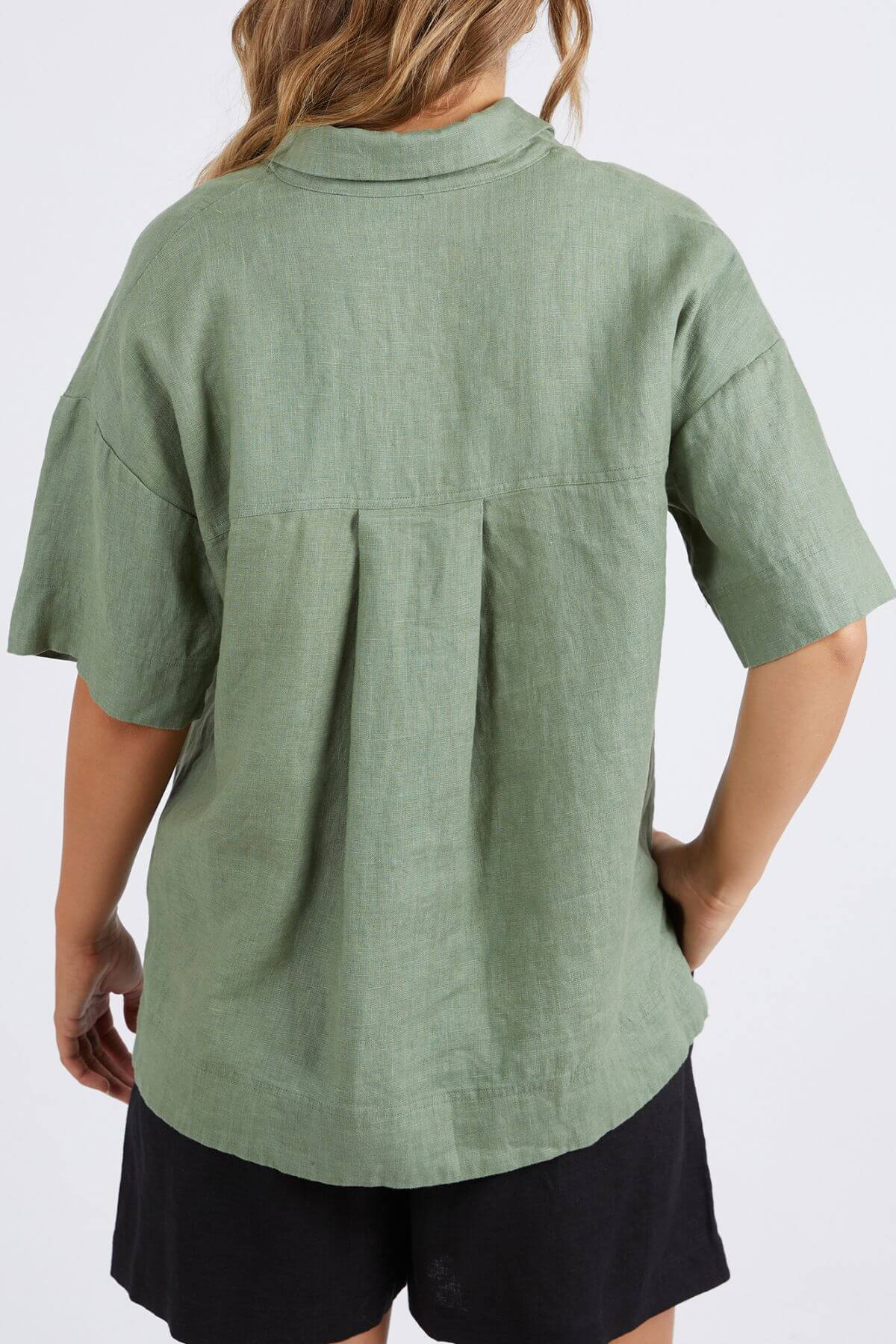 A person wearing a Foxwood Aubrey Shirt in light green and black shorts is shown from the back. The loose-fitting, short-sleeved linen shirt has pleats near the collar, a button-through placket, and a curved hem. With their left hand on their hip, their hair falls over their shoulder.