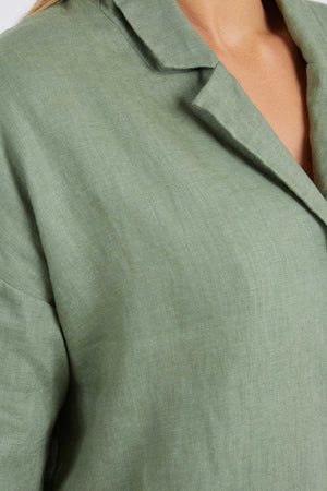 A detailed shot shows the shoulder and chest area of a relaxed-fit Foxwood Aubrey Shirt in light olive green linen, highlighting its button-through placket and fabric texture.
