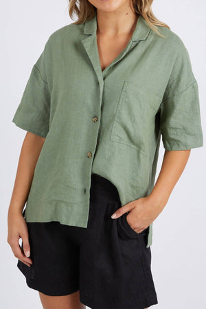 A woman wears the Foxwood Aubrey Shirt, a loose-fitting, light green linen top with short sleeves, a button-through placket, and a left-side pocket. She pairs it with black shorts and rests her left hand in her shorts pocket against a plain background.