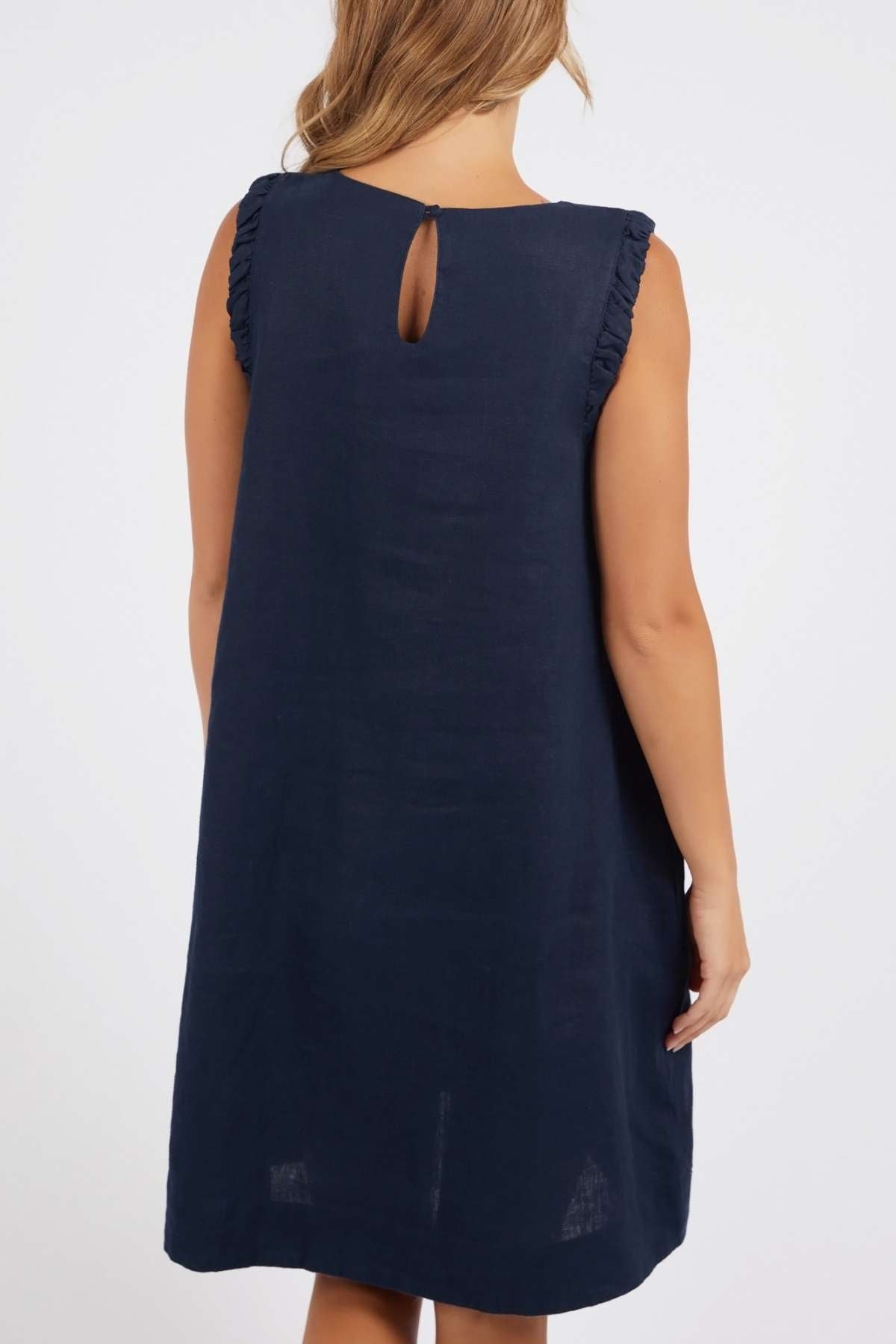 A woman wears the Foxwood Bastia Dress, a navy blue sleeveless, knee-length A-line dress with ruffled shoulder details, shown from the back against a plain background.