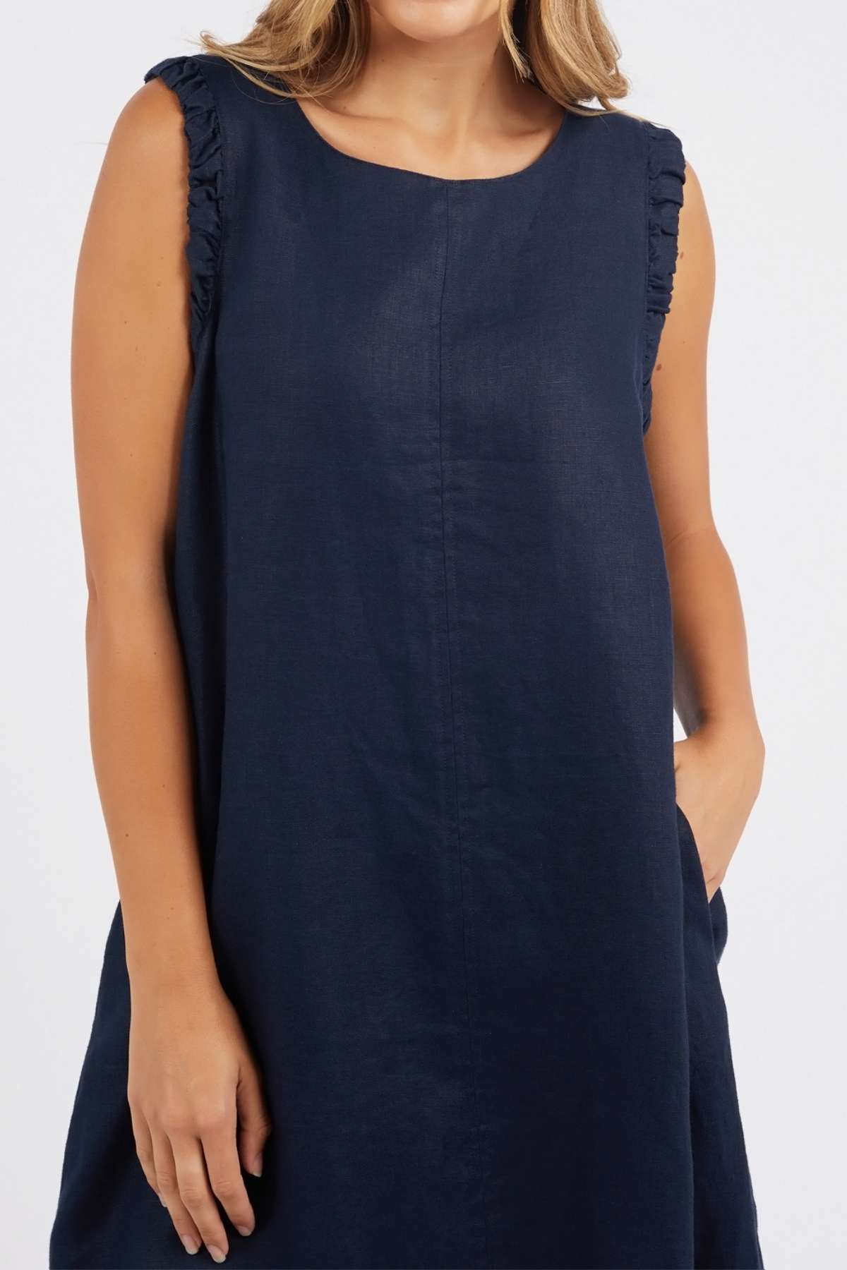 A woman stands against a plain light background, wearing the Foxwood Bastia Dress—a sleeveless, loose-fitting navy blue A-line style with ruffled shoulder detailing and one hand in her pocket.