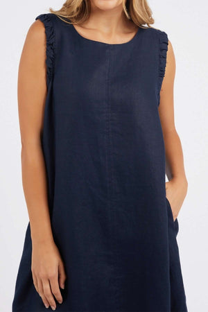A woman stands against a plain light background, wearing the Foxwood Bastia Dress—a sleeveless, loose-fitting navy blue A-line style with ruffled shoulder detailing and one hand in her pocket.