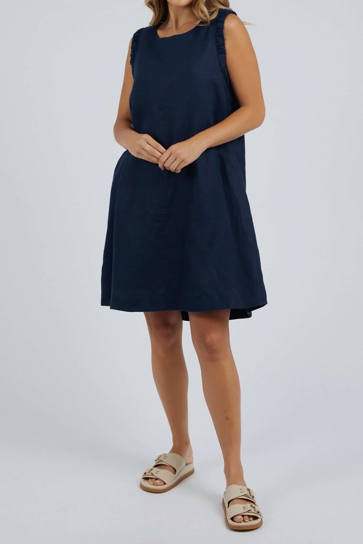 A woman stands against a plain light background, hands lightly clasped in front of her, wearing the Foxwood Bastia Dress—sleeveless, loose-fitting, and navy blue linen—with beige sandals.