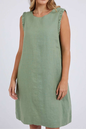 The woman elegantly poses against a plain backdrop, highlighting the Foxwood Bastia Dress, a sleeveless green A-line piece with knee-length and charming ruffled shoulders that enhance its sophisticated allure.