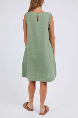 A person, back facing the camera, wears a sleeveless green Foxwood Bastia Dress with ruffled shoulders and a keyhole opening. This elegant A-line dress is paired with tan sandals against a plain white background.