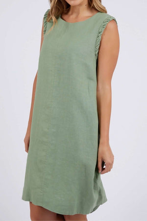 A person stands against a neutral background, wearing the Foxwood Bastia Dress—a sleeveless, knee-length green linen dress with ruffled shoulders and a simple, relaxed fit. Their arms are relaxed by their side.